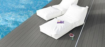 Elegante Poolterrasse mit dunklem WPC-Belag, zwei weißen Lounge-Liegen und ruhiger Wasserreflexion.