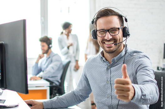 Freundlicher Kundenberater mit Headset sitzt an einem Schreibtisch mit Computer und zeigt den Daumen hoch.