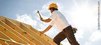 Dachdecker mit gelbem Schutzhelm beim Befestigen von Konstruktionsvollholz (KVH) auf einem Dachstuhl unter strahlendem Himmel.