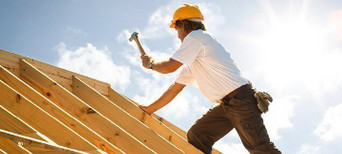 Dachdecker mit gelbem Schutzhelm beim Befestigen von Konstruktionsvollholz (KVH) auf einem Dachstuhl unter strahlendem Himmel.