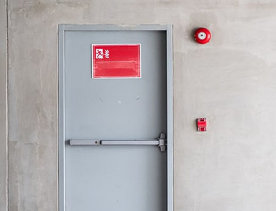 Graue Brandschutztür mit Fluchtwegsymbol und roter Alarmglocke.